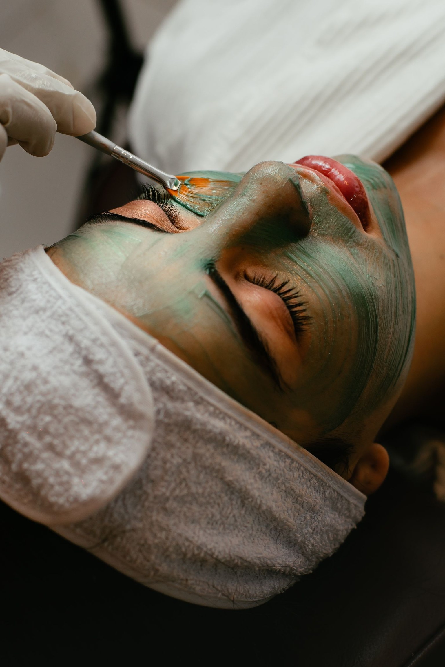 Une esthéticienne applique un masque vert au pinceau sur le visage d’une personne allongée, les yeux fermés.