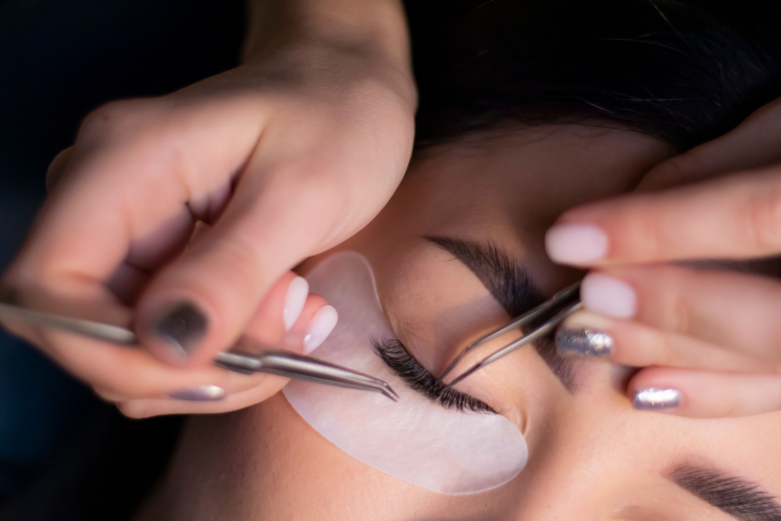 Une professionnelle utilise deux pinces de précision pour poser des extensions sur les cils d’une cliente aux yeux fermés.