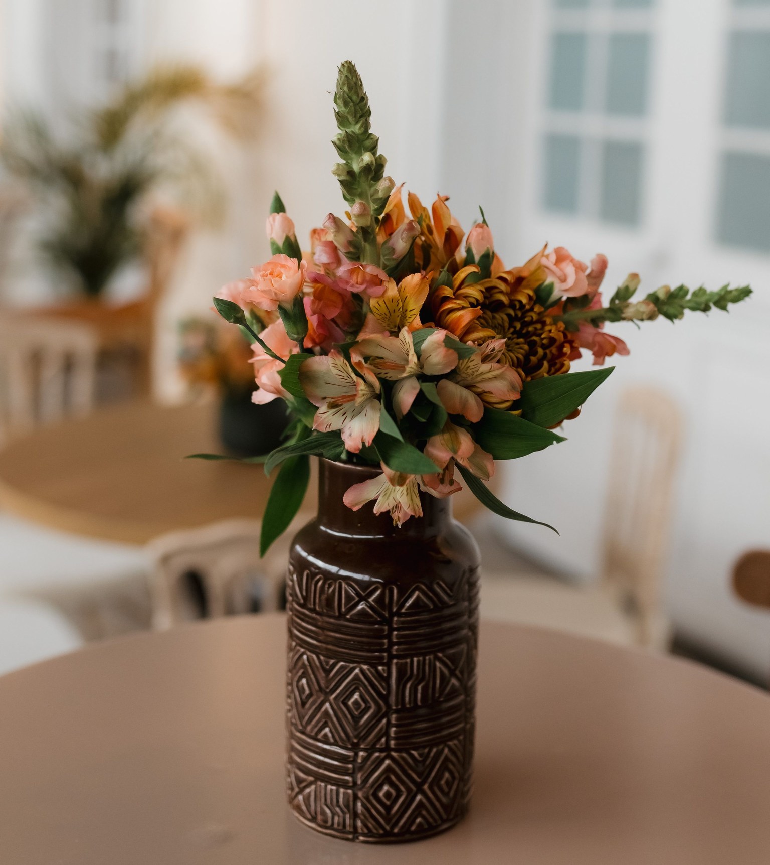 Fleurs roses et orangées dans un vase sombre à motifs, posé sur une table.