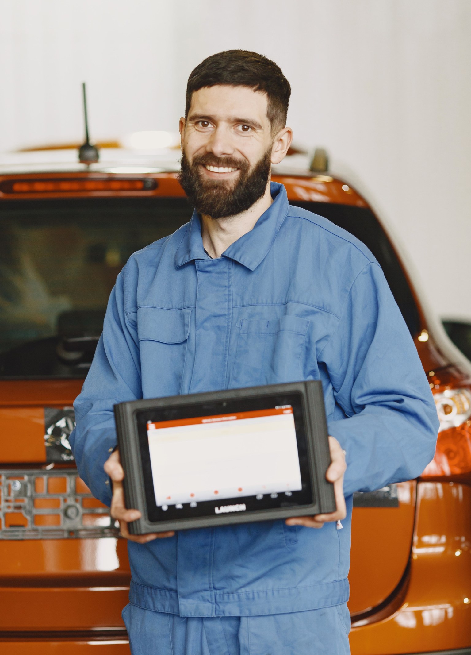 Technicien en combinaison bleue présentant une tablette de diagnostic automobile devant une voiture.