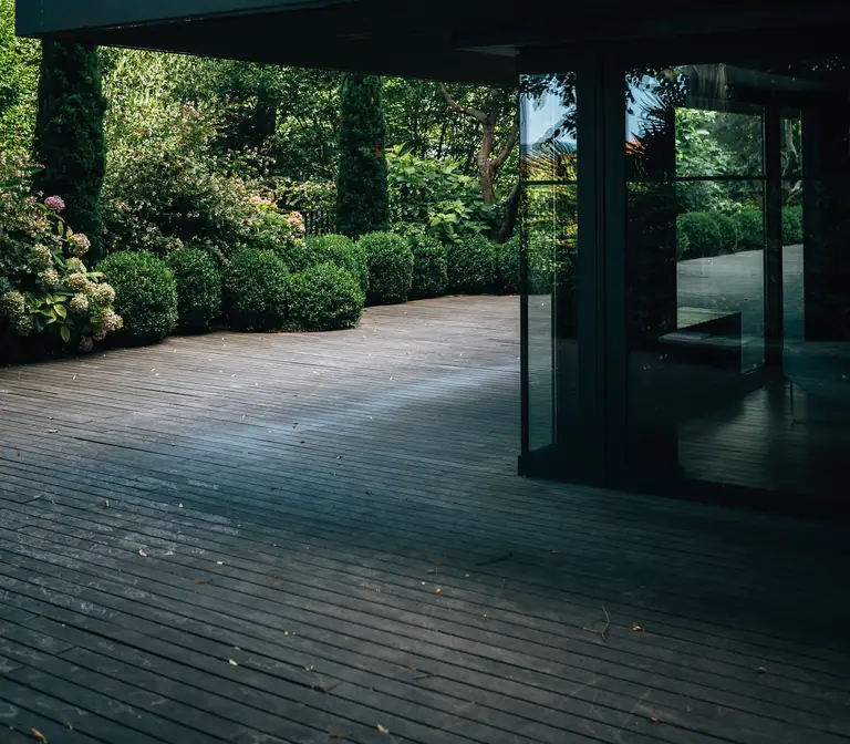 Une terrasse en lames de bois longe une façade vitrée et des arbustes taillés dans un jardin.