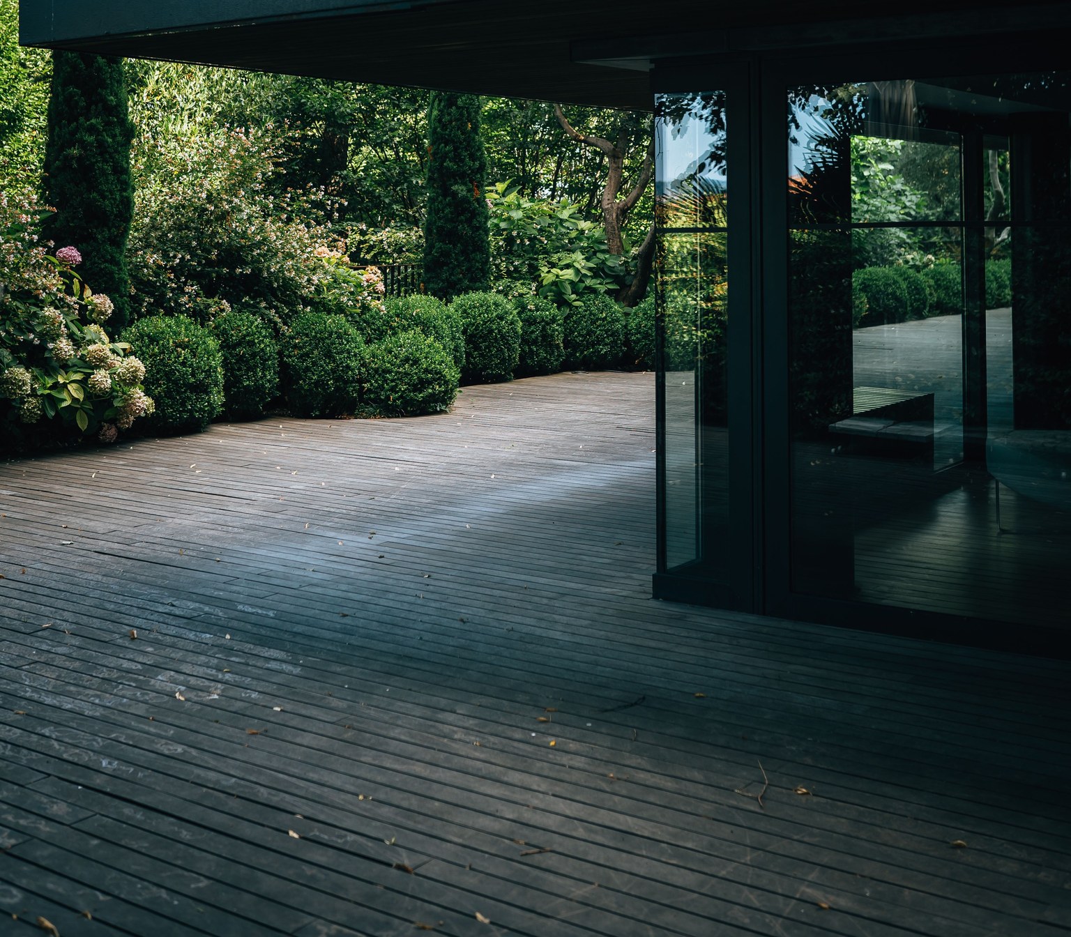 Une terrasse en lames de bois longe une façade vitrée et des arbustes taillés dans un jardin.